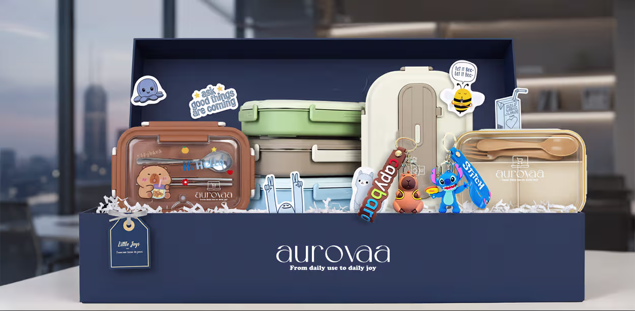 Aurovaa 'Little Joys' gift box filled with cute pastel lunch boxes, bento containers, wooden cutlery, Stitch and animal-themed keychains, fun stickers ('Let it Bee', 'Hi Hello'), and playful accessories on white shredded filler — kawaii daily-use joy set in navy blue packaging.